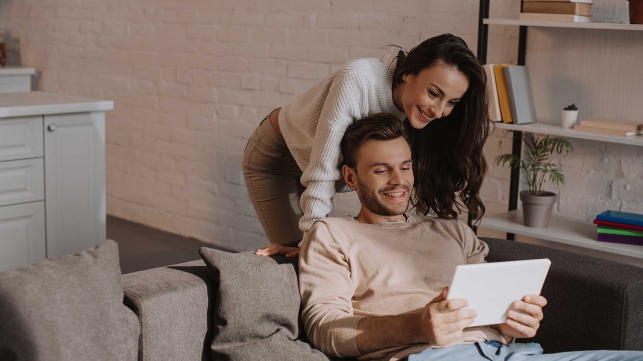 Ein Mann mit einem Tablett auf dem Sofa und die Frau steht hinter ihm und schaut sich das Tablet an